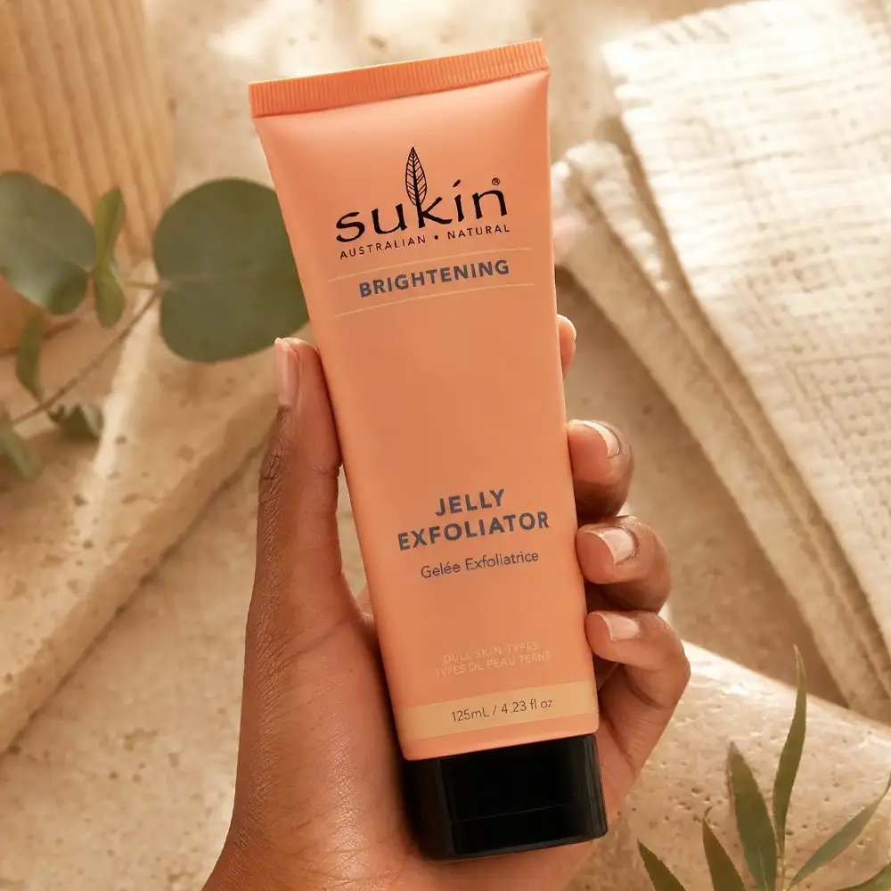 Hand holding Sukin Brightening Jelly Exfoliator against warm stone and botanical details